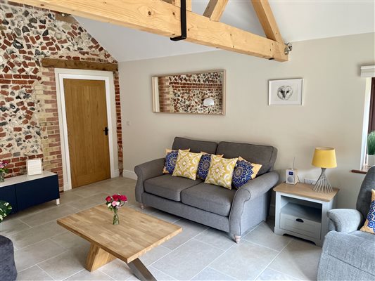 Grey sofa on your right with yellow cushions. Dark grey chair on the left with bright coloured cushions.  Original flint wall on the left with a wooden door to the right. In the centre is a wooden coffee table with fresh red flowers on it.